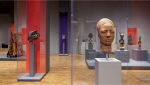 Image of a museum gallery with grey plinths containg objects the closest object is a carved wooden head, possibly African.