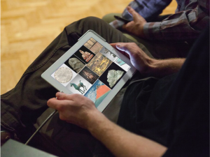 Image of a person sitting with a tablet computer on their lap with images of objects in squares on the screen. You can only see the persons torso and legs in the image.