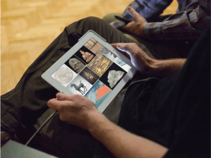 Image of a person sitting with a tablet computer on their lap with images of objects in squares on the screen. You can only see the persons torso and legs in the image.