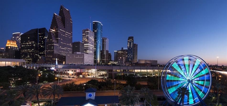 An image of the Houston skyline at sunset with skyscrapers in the background and a ferris wheel lit with blue lights in the foreground