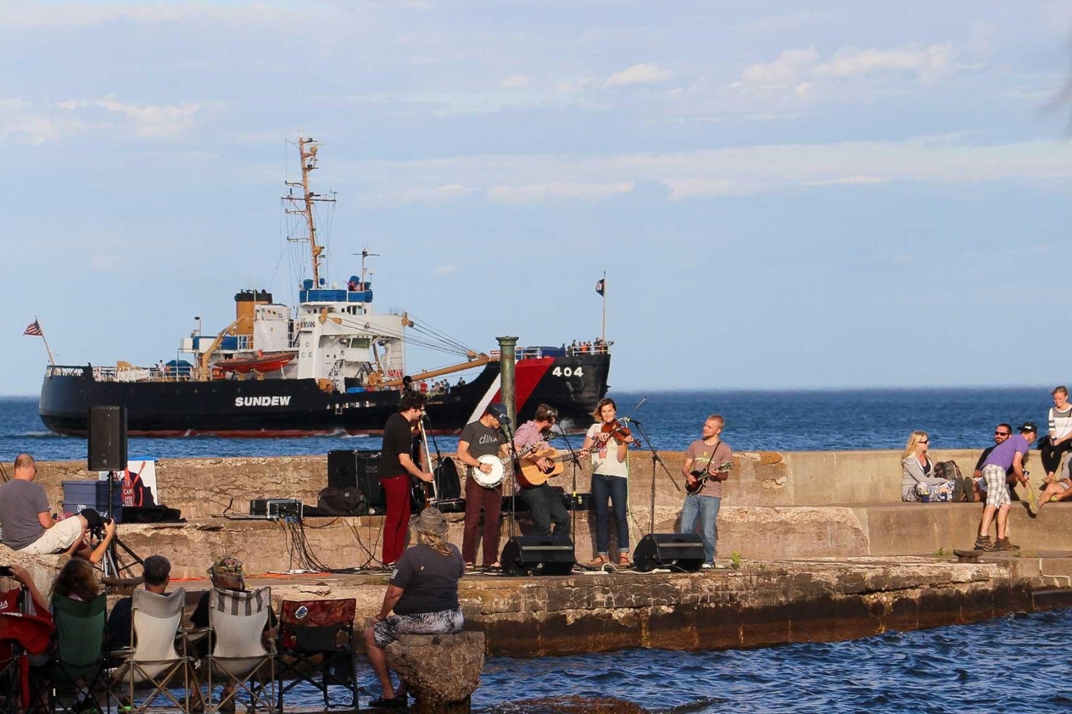 A group of people play instruments on a concrete pier in the middle of the water. A large ship is seen in the background and a small group of peopel sit in fabric folding chairs in the foreground. 