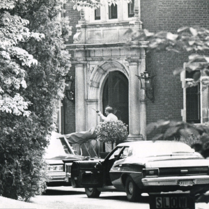 Black and white image of police cars parked in front of the mansion with one gentleman standing near the front door gesturing with what appears to be a walkie talkie in his hand. 