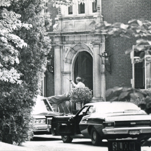 Black and white image of police cars parked in front of the mansion with one gentleman standing near the front door gesturing with what appears to be a walkie talkie in his hand.