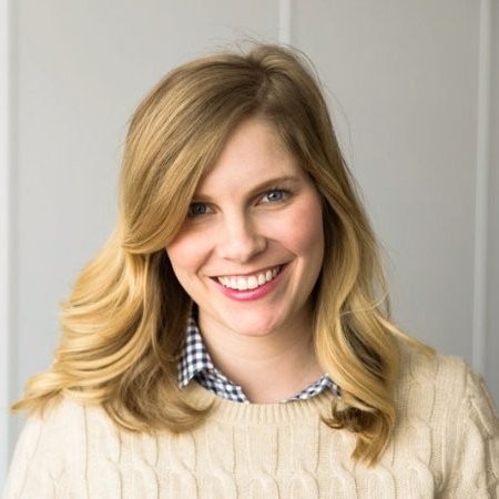 A white woman with shoulder length blond hair smiles at the camera wearing a cream colored cable knit sweater.