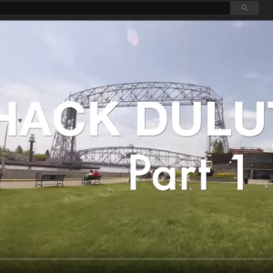 Image of a video screen with the words Hack Duluth Part 1 overlayed on top of an image of a bridge and park like area with people and dogs milling about. 