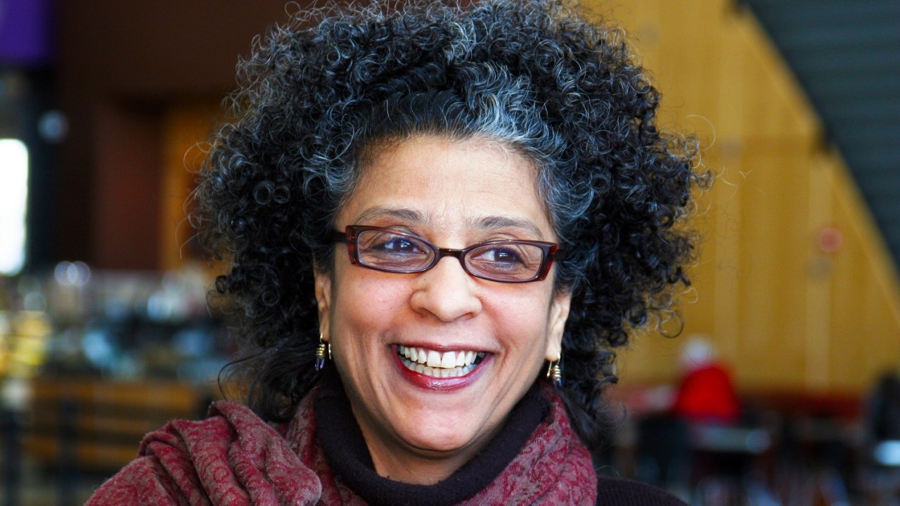 A woman smiling looking off into the distance with short curly black hair with bit of grey wearing red rimmed glasses and dangling earrings with a pink scarf around her neck.