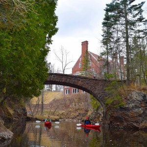 Image of two people in kayaks traveling toward the camera floating under a stone bridge with rock walls on either side, heavy plant growth to the left and Glensheen mansion visible behind them.