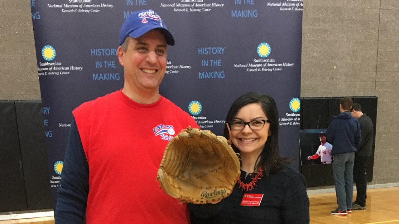 A man and woman face the camera smiling wearing baseball gear the man holds a baseball glove and hat.
