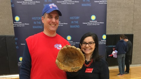 A man and woman face the camera smiling wearing baseball gear the man holds a baseball glove and hat.