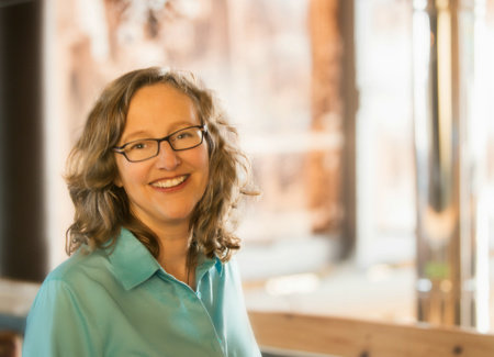 Image of a Lucie sitting near an open window which is fuzzy in the background. She is wearing a pale green top and dark rimmed glasses. She has shoulder length wavy dirty blond hair.