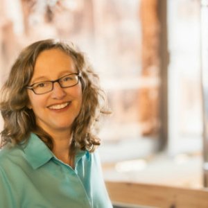 Image of a Lucie sitting near an open window which is fuzzy in the background. She is wearing a pale green top and dark rimmed glasses. She has shoulder length wavy dirty blond hair.