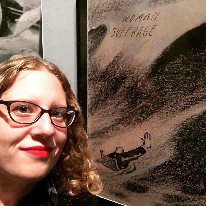 Selfie of a white woman with blond curly hair wearing dark rimmed glasses standing next to a framed Woman's Suffrage sketch.