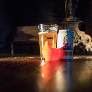 Image taken at floor level of a glass with an amber colored liquid, a can of Ballast Point beer, and a sippy cup lid.