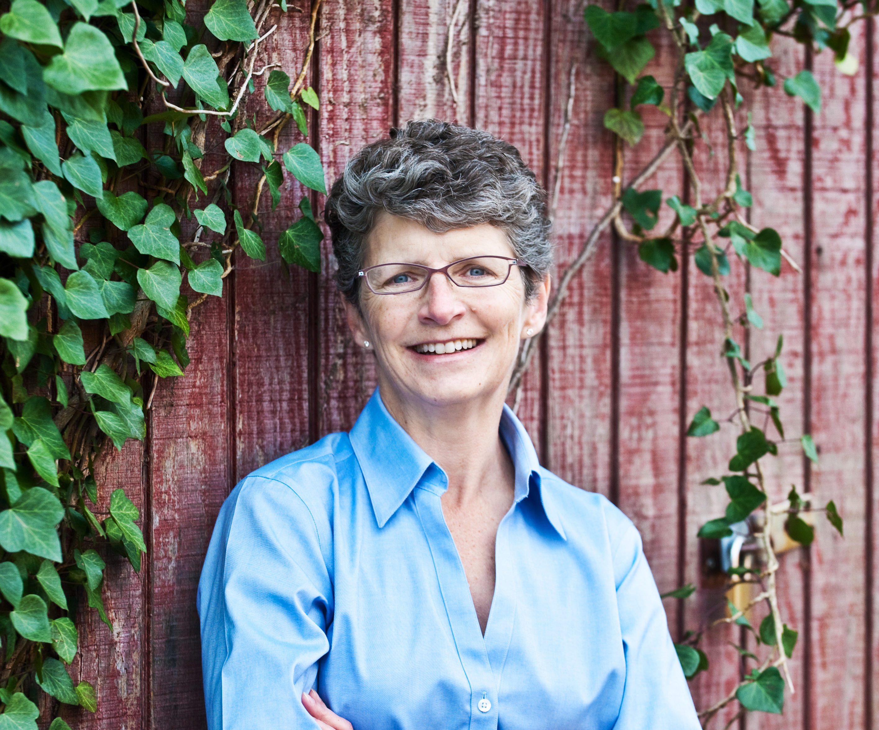 Headshot of a woman standing in front of a wooden wall with green leafy ivy hanging from above.