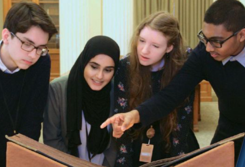 Four teenagers look at a manuscript with a boy on the right pointing at the book below them.