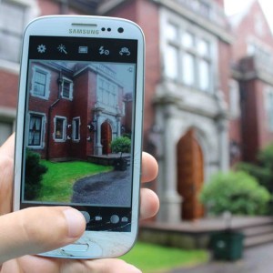 Image of someone holding up their cell phone to take a picture of the outside of Glensheen mansion. You can see the house blurry in the background and clear on the phone screen in the image. 