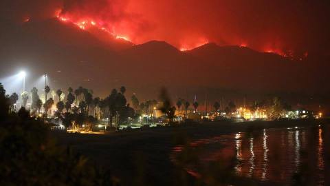 Image shows a beach area with a populated area and lights and in the background a mountain area with a red fire line all along it.