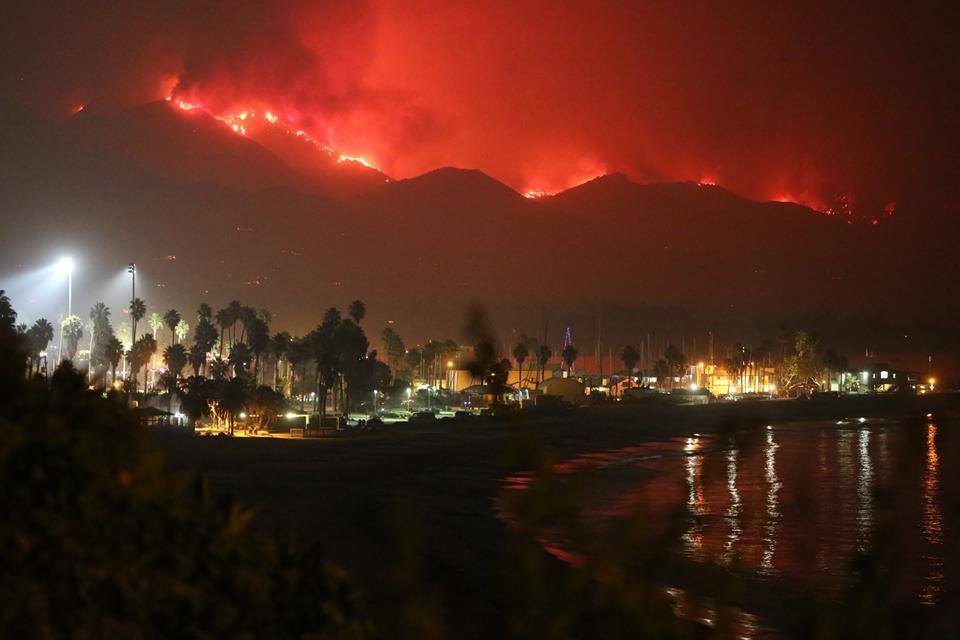 Image shows a beach area with a populated area and lights and in the background a mountain area with a red fire line all along it.