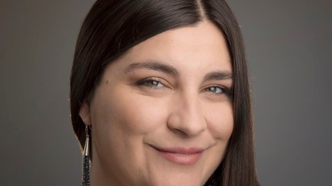 Headshot of a woman standing in front of a dark grey backdrop with her long dark brown hair pulled over her right shoulder. She is wearing a veritcal black and white striped top and has long dangling earrings on.