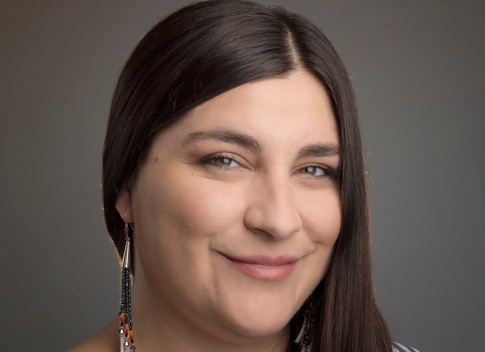 Headshot of a woman standing in front of a dark grey backdrop with her long dark brown hair pulled over her right shoulder. She is wearing a veritcal black and white striped top and has long dangling earrings on.