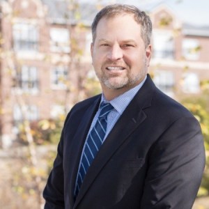 A man stands outside smiling at the camera wearing a dark colored suit and blue and white striped tie.