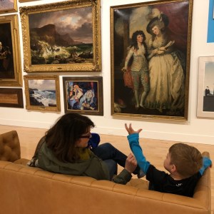 A mother and her son sit on a couch facing a grouping of paintings on the wall opposite.
