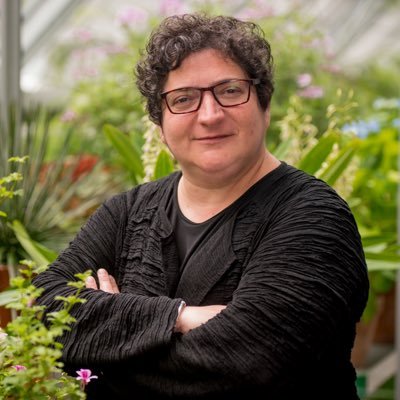 A woman stands in a greenhouse wearing a black blouse with her arms folded in front of her