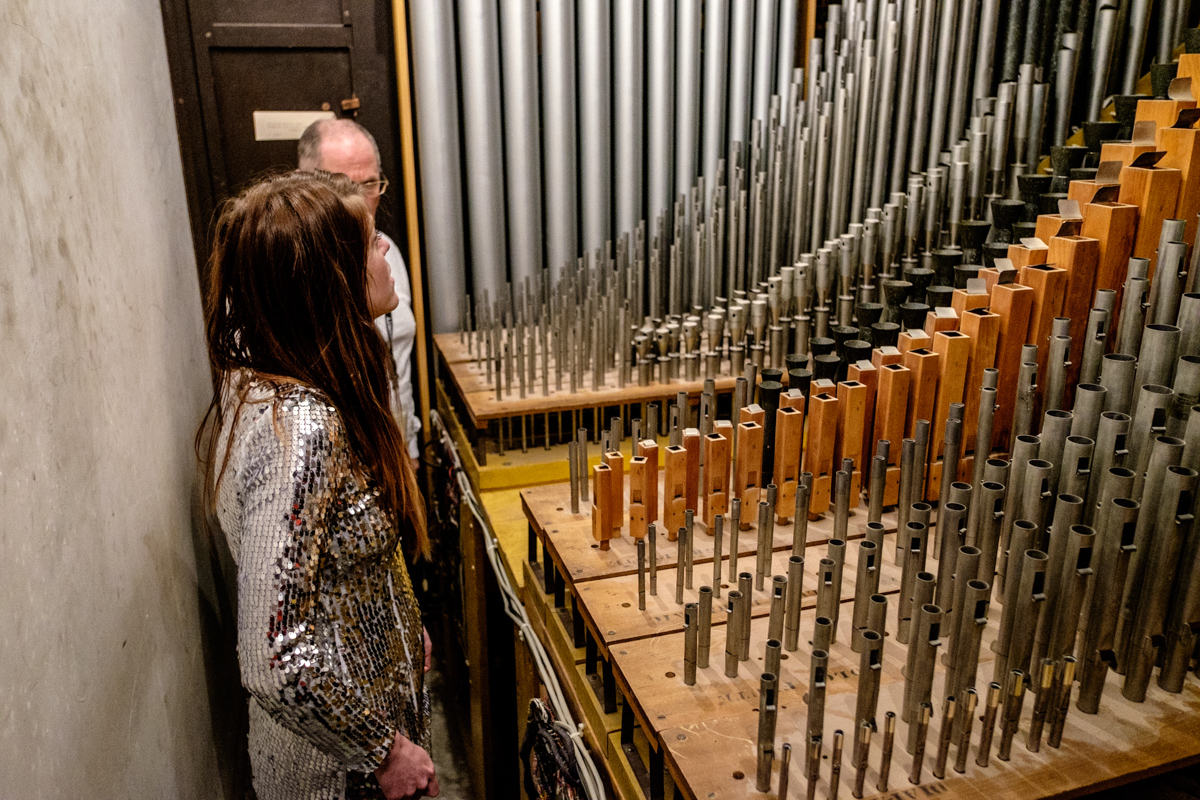 A woman in a sparkly shirt stands next to a set of pipes listening to a guide discuss how the pipe organ is used. 