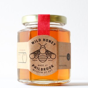 A hexagonal honey jar sits by itself with a bee on the label and a red ribbon around it. 