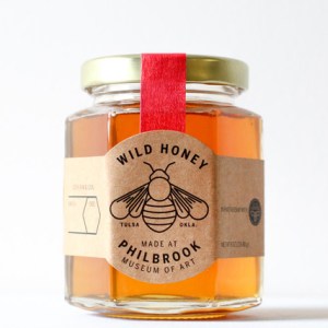 A hexagonal honey jar sits by itself with a bee on the label and a red ribbon around it.