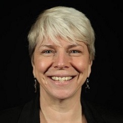 Headshpt of a grey haired woman facing directly into the camera smiling wearing dangling earrings.