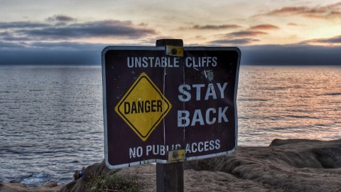 Image of a signpost that reads "Unstable Cliffs, DANGER, Stay Back, No Public Access" in front of an ocean view with sandy cliffs.