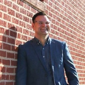 A man stands against a brick wall smiling at the camera wearing wire rimmed glasses and a blue blazer with a blue three button shirt.