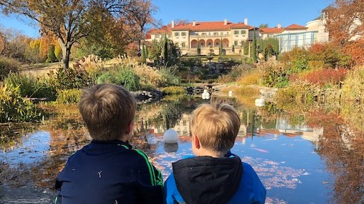 Two young boys sit on a rock and look across a pond to a large Spanish style mansion on a hill.