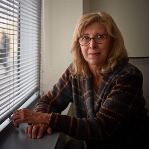 Michelle Smith sits next to a window with blinds on it with her hands crossed on the window sill. She has shoulder length blond hair and is wearing red rimmed glasses, she is also wearing a plaid jacket.