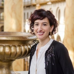 A woman stands next to a gilded urn in front of large indoor columns smiling at the camera. She has short curly borwn hair and is wearing a white shirt with a lacy black sweater.