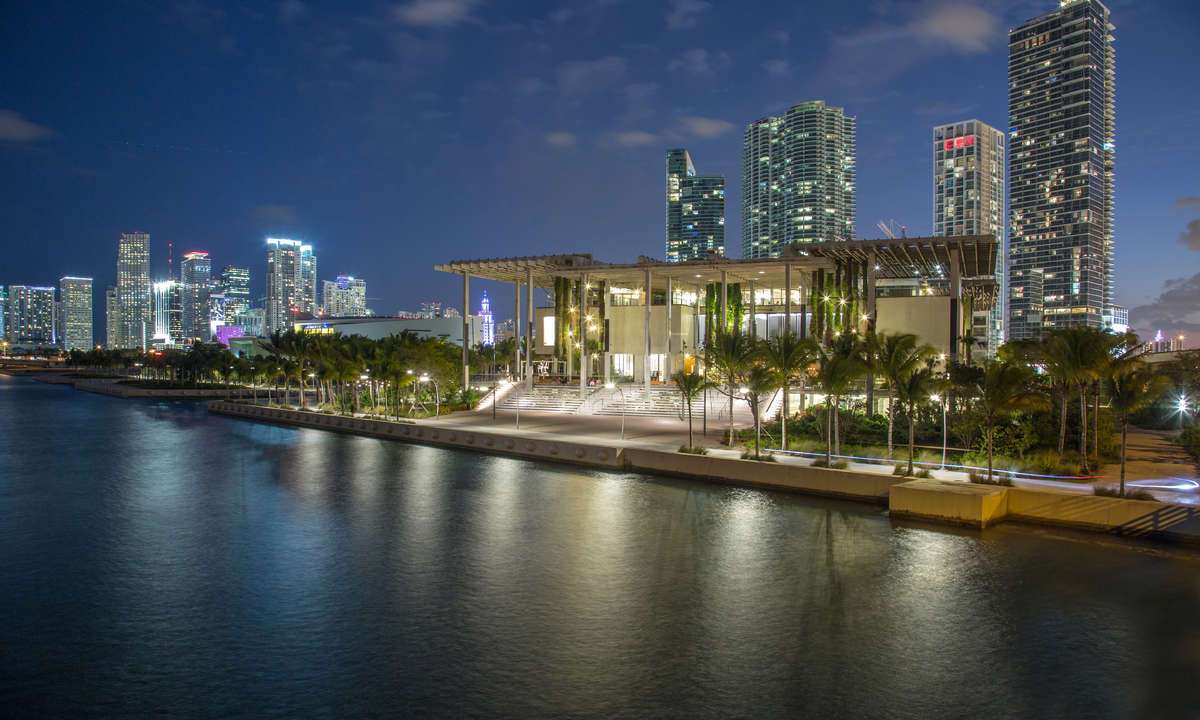 Photo of a contemporary building (the Perez Art Museum Miami) from the water. It's dusk and the Miami skyline is visible in the background.