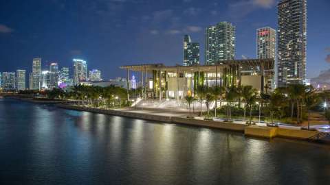 Photo of a contemporary building (the Perez Art Museum Miami) from the water. It's dusk and the Miami skyline is visible in the background.