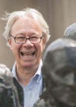 A white haired white man wearing dark wire rimmed glasses smiles laughingly at the camera standing behind several copper colored busts.