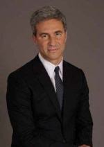 A white man stands in front of a grey backdrop wearing a dark suit and tie.