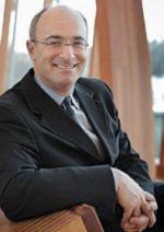 A white man sits in a comfortable chair in front of curtained windows turning to look at the camera wearing wire rimmed glasses and dark suit.