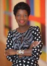 A black woman stands in front of a blurred out painting with her arms folded and a smile on her face.