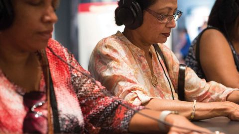 Two women sit at a grey countertop one is clearly wearing headphones both have their hands on the counter looking down.