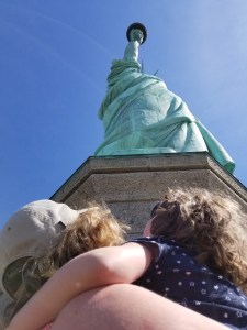 A woman holds a young child in her arms looking up at the Statue of Liberty. We see the backs of their heads, the woman is wearing a ball cap.
