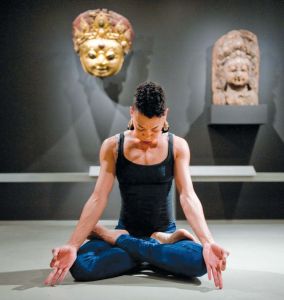 A woman sits in a gallery cross legged with her hands in Om position. 