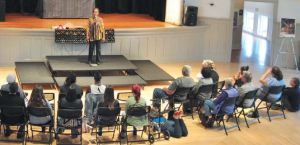 A group of people sit in folding chairs in front of a stage where a woman is standing speaking into a microphone