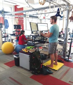 View of an alternative office setting with a white man standing on a yellow foam square and a white woman sitting on a blue yoga ball. Both are looking at their computer screens. 