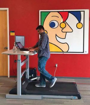 A middle eastern man stands on a treadmill with a desk attached with a joker painting behind him. 