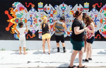 A group of people stand painting an exterior wall with a white woman walking by. 
