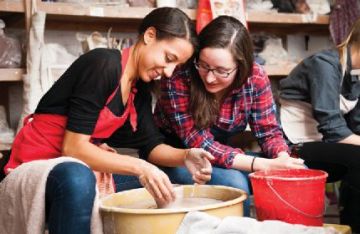 Two women sit next to a pottery wheel watching a piece of clay being formed by one of the women.